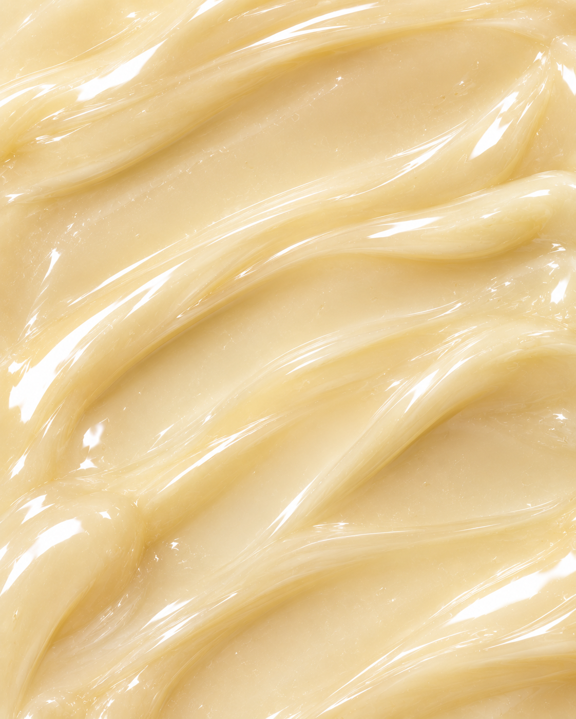 A close-up texture shot of a thick, glossy cream or balm with smooth, swirled ridges and warm beige-yellow tones.