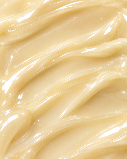 A close-up texture shot of a thick, glossy cream or balm with smooth, swirled ridges and warm beige-yellow tones.